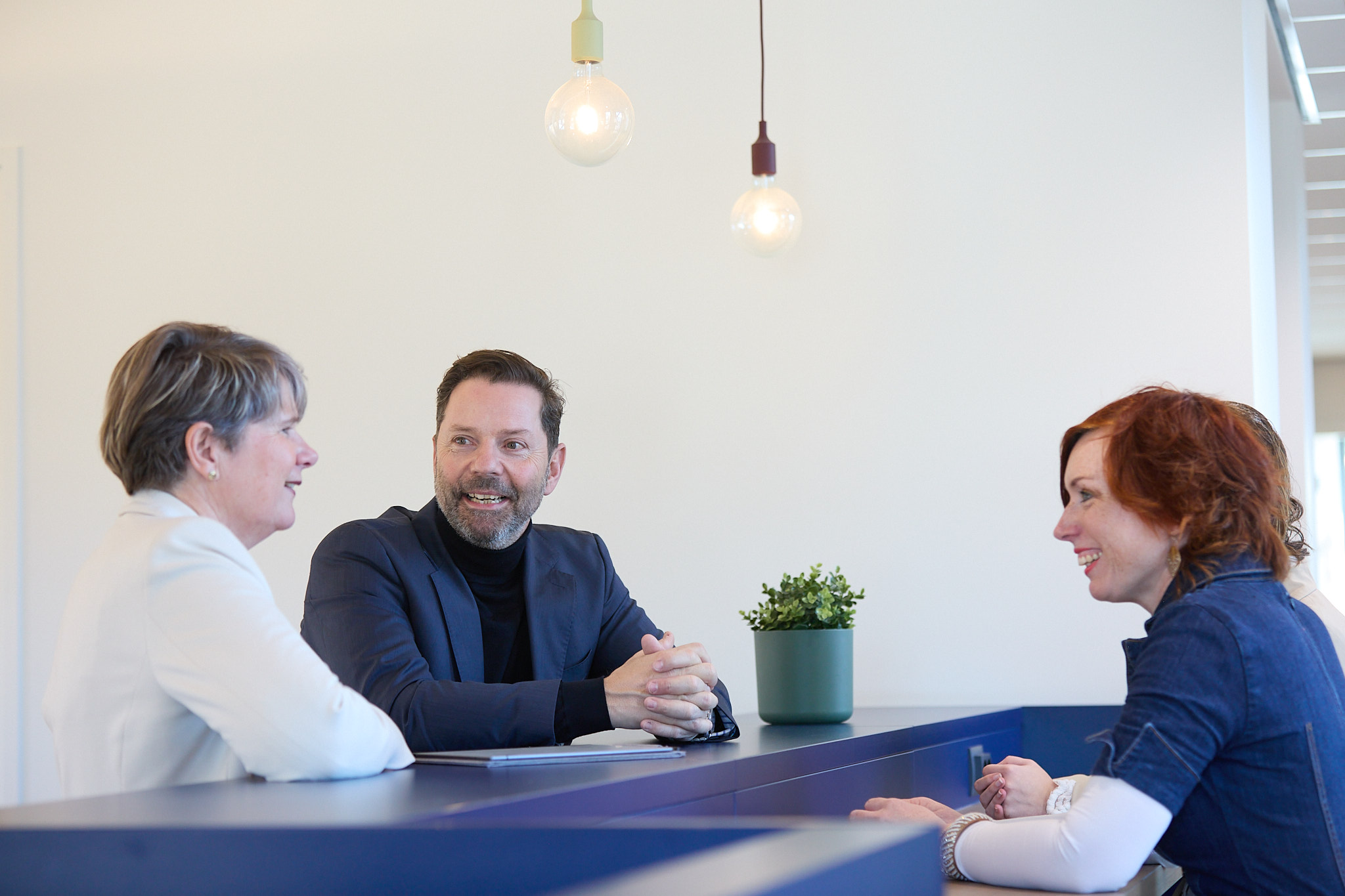 Twee vrouwen en man voeren kennismakingsgesprek aan balie met hanglampen op achtergrond.