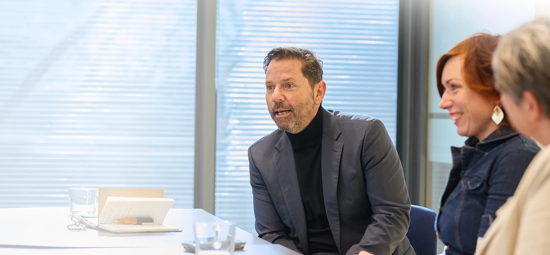 Arbeidsdeskundige in donker colbert bespreekt casus met twee lachende collega’s aan vergadertafel.